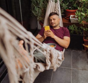 Young man relaxing on the terrace of his apartment, lying in a hammock and using smart phone