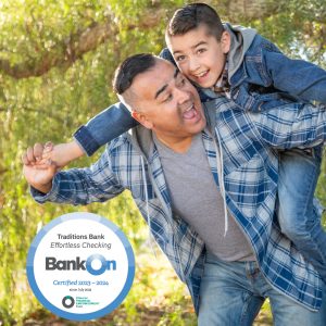 Dad holding his son on his back outside by a tree with the Traditions Bank Effortless Checking Bank On certification seal for 2023 - 2024