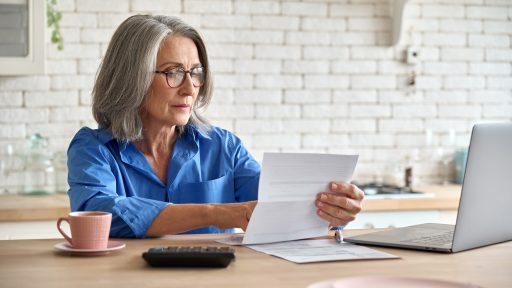 senior woman working from home, looking at paperwork with laptop open