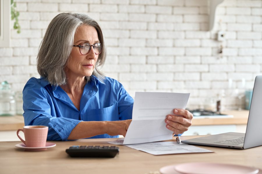 senior woman working from home, looking at paperwork with laptop open