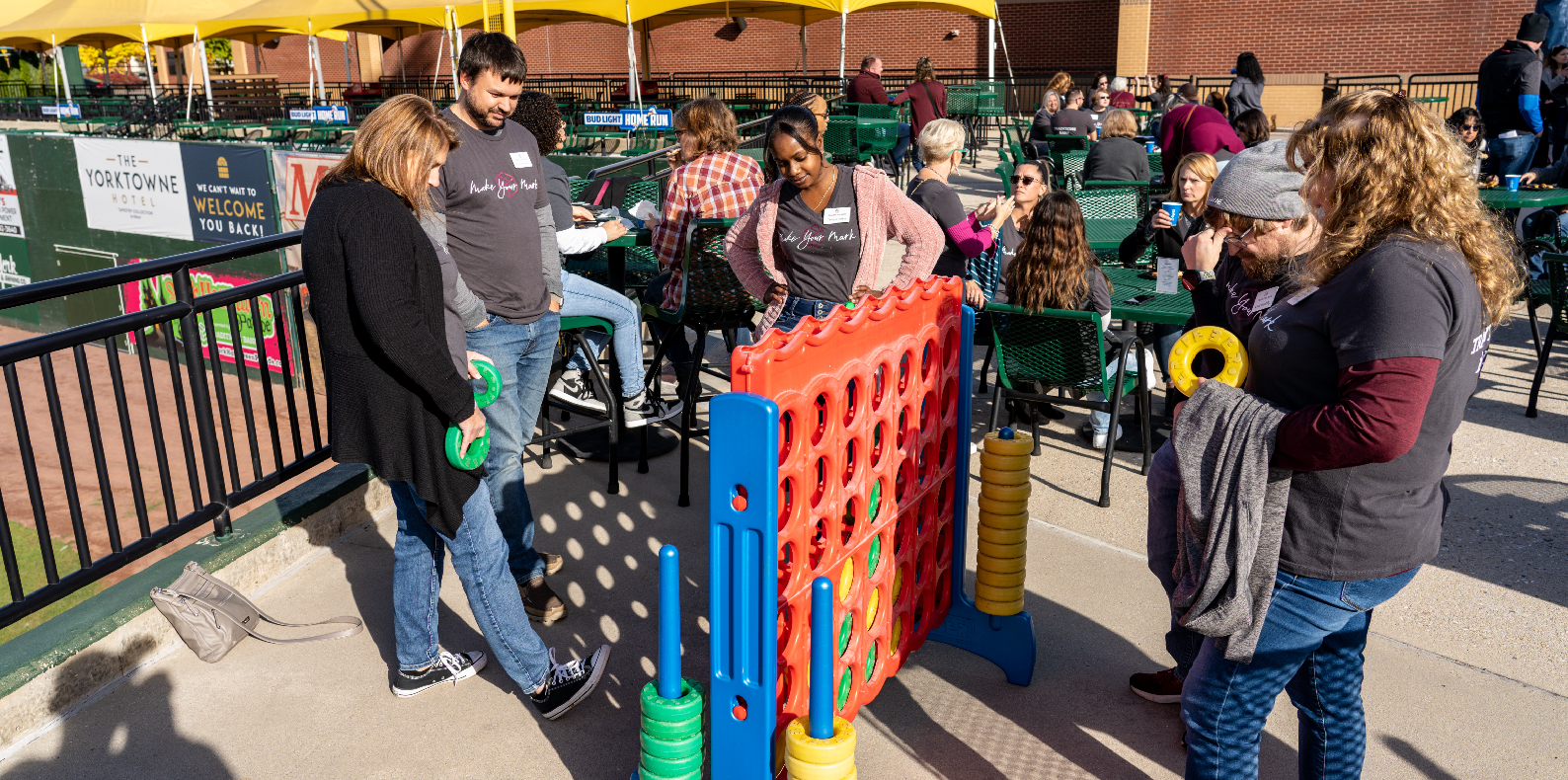 group of male and female coworkers playing a game of giant connect four at a company party in a baseball stadium