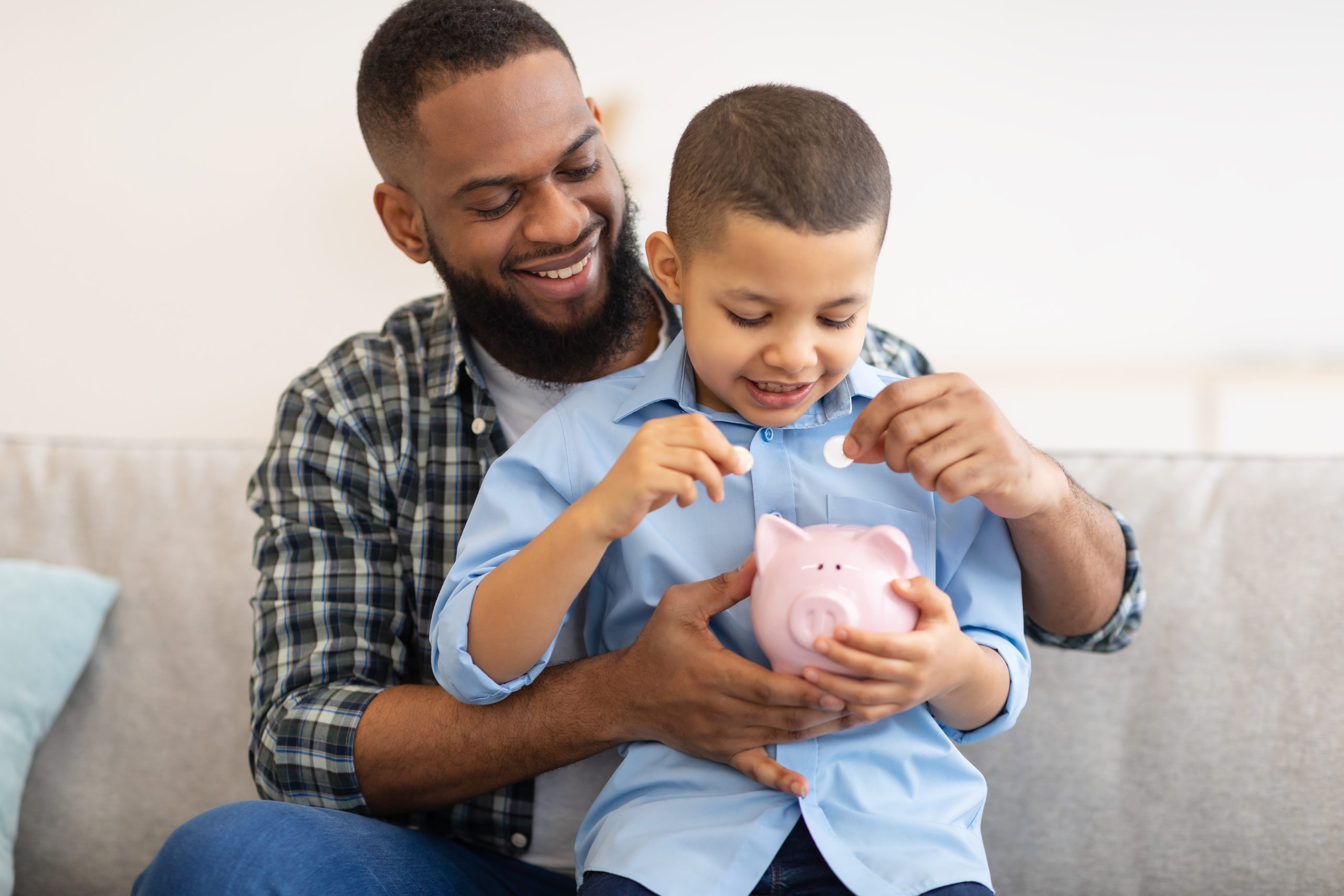 Dad And His Toddler Son Putting Coin In Piggybank Sitting On Couch At Home.