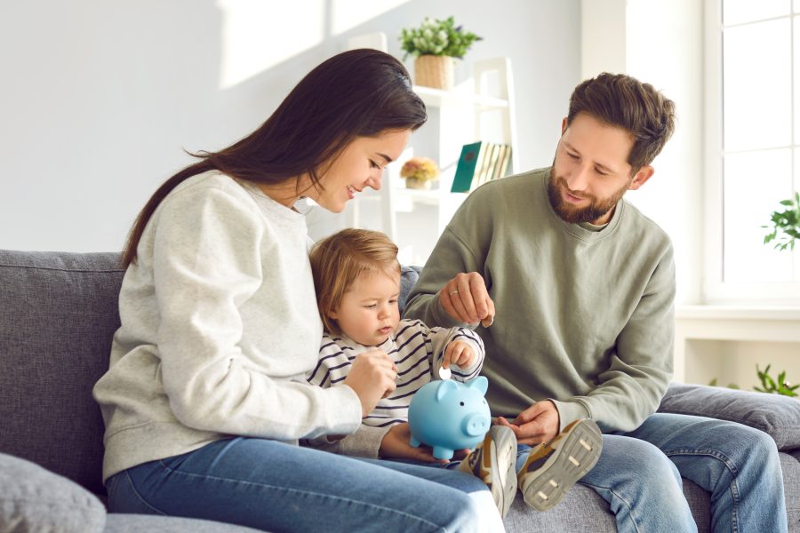 Caring parents teaching toddler baby how to save money. Mom, dad and kid holding piggy bank sitting on sofa at home.