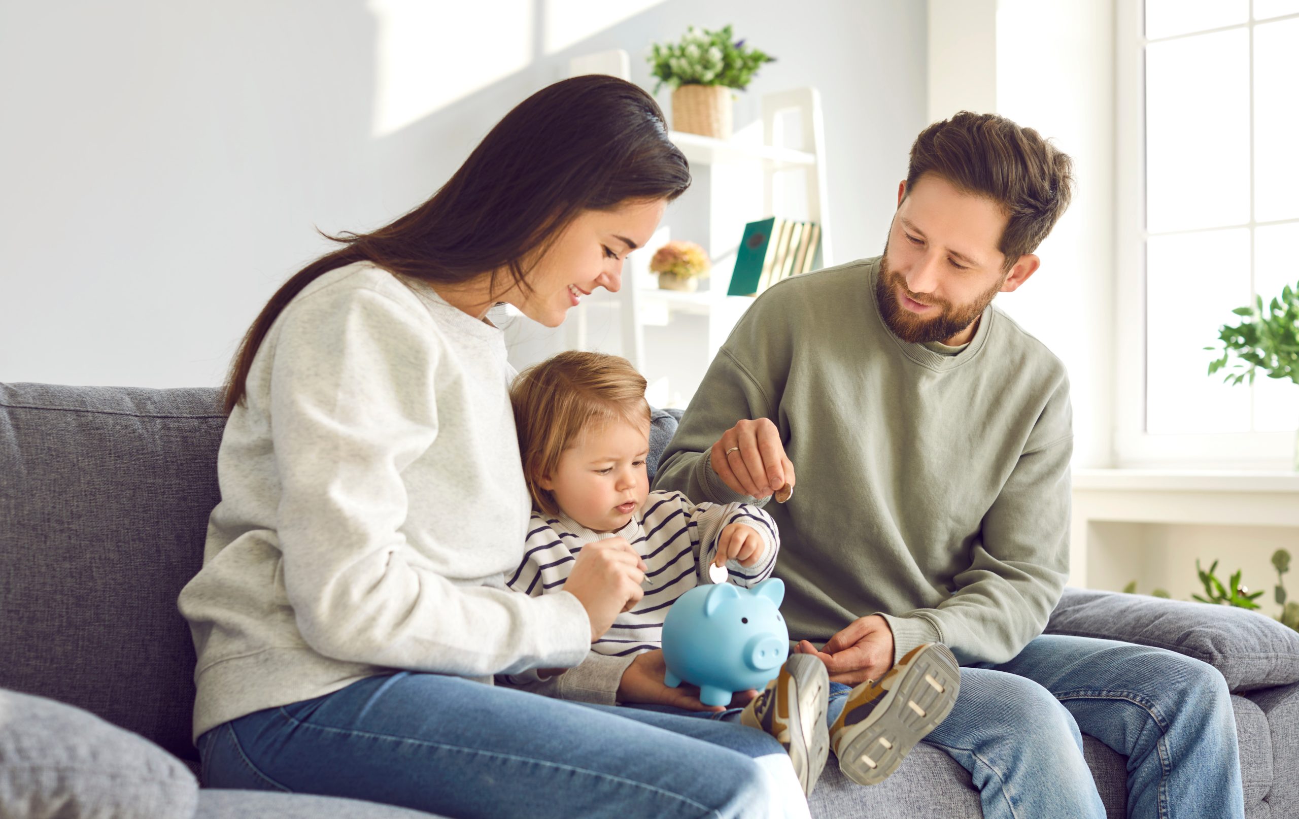 Caring parents teaching toddler baby how to save money. Mom, dad and kid holding piggy bank sitting on sofa at home.